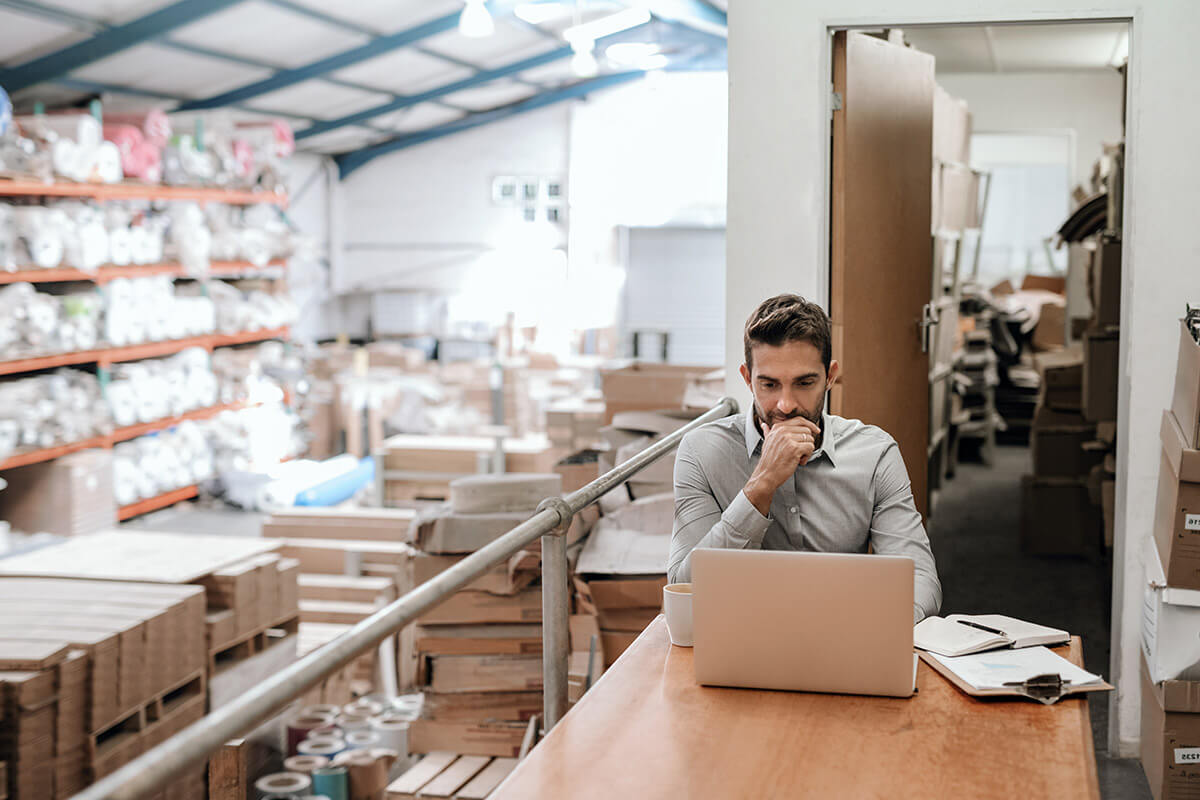 manager working on a laptop while sitting at a desk in a warehouse with shelves of stock in the background